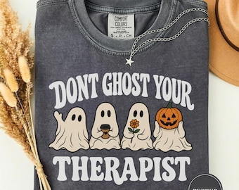 May include: A charcoal gray t-shirt with a vintage wash, featuring the white text DONT GHOST YOUR THERAPIST. The graphic includes four white ghosts: one waving, one holding a coffee cup, one holding a flower, and one holding a jack-o-lantern.