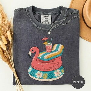 May include: A charcoal gray crew neck t-shirt featuring a graphic of a pink flamingo pool float stacked on a teal floral inner tube with a striped lounge chair and a tropical drink. The shirt has a Comfort Colors label at the inner neckline.