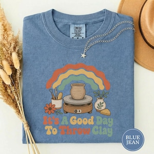May include: A blue short-sleeve t-shirt featuring a graphic of a pottery wheel, a clay pot, tools, and a plant under a rainbow. The text reads It's A Good Day To Throw Clay. The shirt has a crew neckline and a Comfort Colors brand label at the neck.