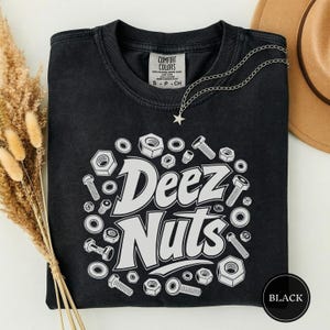 May include: A black short-sleeved t-shirt featuring a white graphic print of the text "Deez Nuts" surrounded by various illustrations of metal nuts and bolts. The shirt has a crew neckline and a Comfort Colors brand label on the inside collar.