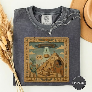 Könnte beinhalten: Ein anthrazitfarbenes T-Shirt mit Rundhalsausschnitt und einem Grafikdruck, der einen Außerirdischen und eine altägyptische Person vor einer Pyramide mit einem darüber schwebenden UFO zeigt. Das Motiv ist von einer dekorativen Bordüre umgeben.