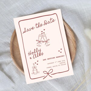 May include: A rectangular Save the Date card with a cream background and dark red line art. It features two champagne flutes, a bottle, and hearts. The text reads: Save the Date, Holly & Alex ARE GETTING MARRIED! SATURDAY 1ST JUNE 2024 CARDIFF.