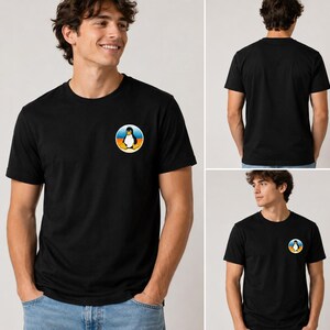 以下が含まれることがあります： 左胸に小さな円形のグラフィックが施された黒の半袖Tシャツ。グラフィックには、黄色いクチバシと足を持つ白黒のペンギンが描かれており、背景には青い空と黄色い地平線がデザインされています。