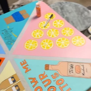 Op de afbeelding: Een driehoekig speeltafeloppervlak geschilderd in secties met lichtblauwe, roze en gele panelen. Eén sectie bevat een illustratie van een Tito's wodkafles met de tekst SHOW ME YOUR TITO'S, terwijl een andere sectie gele citroenschijfjes toont.