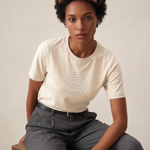 May include: A cream-colored short-sleeve t-shirt featuring a black geometric spiral line art graphic on the chest, paired with charcoal gray pleated trousers and a thin black belt with a silver buckle.