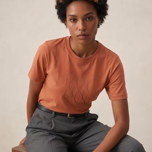 May include: A short-sleeved crew neck t-shirt in a solid terracotta orange color featuring a central graphic design of thin, dark, vertical wavy lines. The shirt is paired with charcoal gray trousers and a thin black belt.