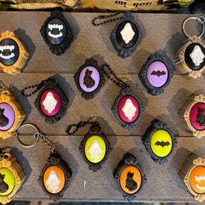 May include: A collection of oval-shaped keychains and charms featuring ornate frames in gold, black, and silver. The centers display raised motifs including white ghosts, black cats, bats, and vampire fangs against colorful backgrounds of orange, purple, pink, and lime green.