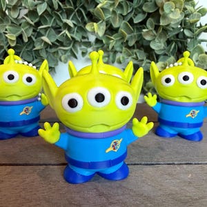 May include: Three small green alien figurines with three eyes, pointed ears, and antennae. Each figure wears a blue space suit with a purple collar and a yellow planet emblem on the chest. The figures have raised arms and are standing in a group.