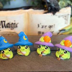 May include: Four small green frog figurines, each wearing a pointed hat. Two frogs wear blue hats with yellow crescent moons, and two wear purple hats with orange pumpkins. The frogs hold small brown wooden-style staffs or broomsticks.