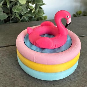 Op de afbeelding: Een klein, rond zwembadspeeltje met drie niveaus en een felroze flamingo-zwemband in het midden. De zwembadstructuur bestaat uit gestapelde ringen in lichtroze, geel en lichtblauw, met een getextureerde, golvende blauwe basis.