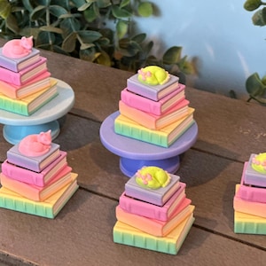 May include: Miniature stacks of four pastel-colored books topped with a small sleeping cat figurine. The books are colored in shades of purple, pink, orange, and green. Some stacks are placed on small, round, solid-colored pedestal stands.