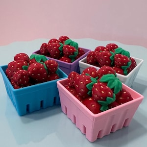 May include: Four square plastic baskets in blue, pink, purple, and white, each filled with small red strawberries featuring green leaves and textured surfaces. The baskets have a slatted design and are arranged together on a light surface.