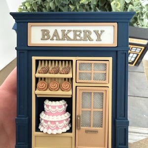 May include: A miniature navy blue bakery storefront model featuring a cream-colored sign with the word BAKERY in raised letters. The display window shows shelves with small brown pastries and a two-tiered pink cake with white frosting details.
