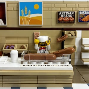 May include: A LEGO bakery playset featuring a minifigure chef in a white uniform behind a counter labelled BREAD - PASTRIES - COFFEE. The display case holds brown bread loaves, while wall signs depict a chef, wheat, and bakery items.