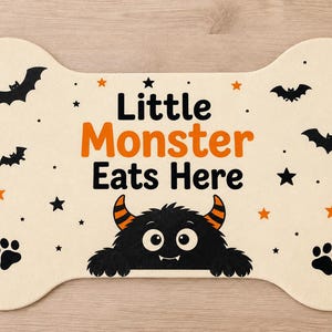 May include: A bone-shaped mat in cream with the text Little Monster Eats Here in black and orange. The design features a black monster with orange-striped horns, black bats, black stars, orange stars, and black paw prints on the surface.