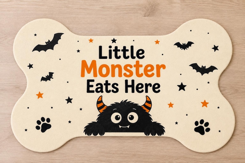 May include: A bone-shaped mat in cream with the text Little Monster Eats Here in black and orange. The design features a black monster with orange-striped horns, black bats, black stars, orange stars, and black paw prints on the surface.
