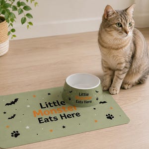 May include: A sage green pet feeding mat and a matching ceramic bowl. The mat features black bat and paw print icons, orange and white stars, and the text "Little Monster Eats Here". The bowl has a white interior and the same text printed on it.