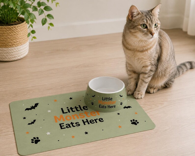 May include: A sage green pet feeding mat and a matching ceramic bowl. The mat features black bat and paw print icons, orange and white stars, and the text "Little Monster Eats Here". The bowl has a white interior and the same text printed on it.