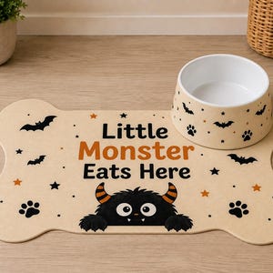 May include: A bone-shaped beige pet feeding mat and a matching ceramic bowl. The mat features the text "Little Monster Eats Here" in black and orange, with black bat, star, and paw print graphics, and a black monster illustration at the bottom.