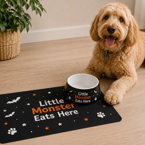 May include: A black pet food bowl and a matching rectangular mat, both featuring the white and orange text "Little Monster Eats Here" alongside white bat, star, and paw print graphics. The bowl is round with a white interior and black exterior.