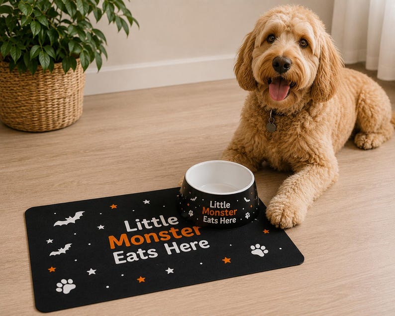May include: A black pet food bowl and a matching rectangular mat, both featuring the white and orange text "Little Monster Eats Here" alongside white bat, star, and paw print graphics. The bowl is round with a white interior and black exterior.