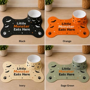 May include: A set of four bone-shaped pet feeding mats and matching bowls in black, orange, ivory, and sage green. Each mat features the text Little Monster Eats Here with bat, star, and paw print patterns. The bowls have a white interior.