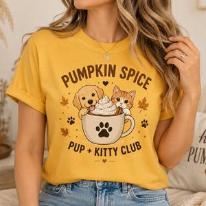 Può includere: Una t-shirt giallo senape con la grafica di un cucciolo di golden retriever e un gattino arancione che spuntano da una tazza piena di panna montata. Il testo recita PUMPKIN SPICE PUP + KITTY CLUB, circondato da piccole foglie d'acero e impronte.
