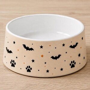 May include: A round ceramic pet bowl with a light beige exterior featuring a repeating pattern of black bats, black paw prints, and small black and orange stars. The interior of the bowl is solid white with a smooth, glossy finish throughout.