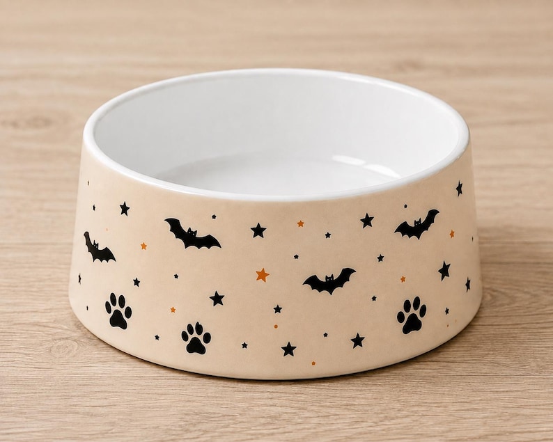 May include: A round ceramic pet bowl with a light beige exterior featuring a repeating pattern of black bats, black paw prints, and small black and orange stars. The interior of the bowl is solid white with a smooth, glossy finish throughout.