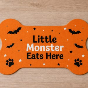 May include: An orange bone-shaped pet mat featuring the black and white text "Little Monster Eats Here". The design includes black bat silhouettes, black paw prints, and small white and black stars scattered across the surface of the mat.