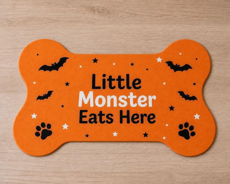 May include: An orange bone-shaped pet mat featuring the black and white text "Little Monster Eats Here". The design includes black bat silhouettes, black paw prints, and small white and black stars scattered across the surface of the mat.