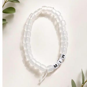 May include: A stretch bracelet composed of translucent, frosted white beads arranged in a circular pattern. Three white square beads with black lettering spelling out the letters M, I, and W are integrated into the design, which is tied with a white cord.