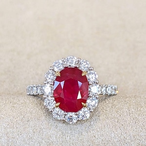 May include: A ring featuring a large, oval-cut, deep red ruby set in a gold prong setting. The center stone is surrounded by a halo of twelve round, brilliant-cut white diamonds. The band is also encrusted with small, round white diamonds.