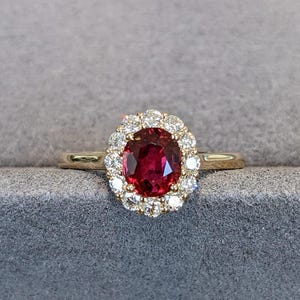 Puede incluir: Un anillo de oro con un rubí central de color rojo intenso y corte ovalado, rodeado por un halo de pequeños diamantes blancos redondos de corte brillante. El aro es liso y pulido, mostrando un diseño clásico con una gema vibrante.