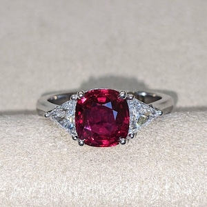 Könnte beinhalten: Ein silberfarbener Ring mit einem großen, tiefroten Edelstein im Kissenschliff in der Mitte. Der zentrale Stein wird auf jeder Seite von einem kleineren, dreieckig geschliffenen, klaren weißen Edelstein flankiert, gefasst in einer polierten Metallschiene.