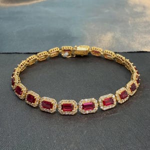 May include: A gold-toned bracelet featuring a series of rectangular, deep red gemstones. Each stone is surrounded by a halo of small, clear, sparkling stones. The bracelet is connected by a metallic clasp mechanism at the top of the circular arrangement.