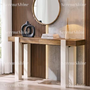 May include: A rectangular wooden console table with two drawers and thick white legs. On the table sits a black abstract sculptural vase, a stack of three books, and a tall, slender beige lamp. A round mirror hangs on the wall above the table.