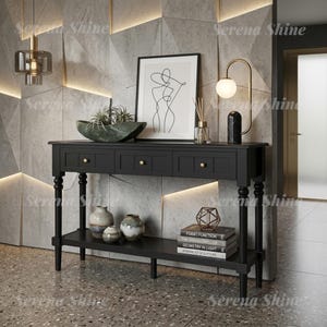 May include: A black wooden console table with three drawers featuring small gold knobs and a lower shelf. The table holds a green bowl with a plant, a framed line art print, a glass lamp, a reed diffuser, three ceramic vases, and a stack of books.
