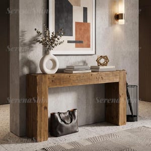 May include: A rectangular wooden console table with a natural grain finish. On top sits a white donut-shaped vase with dried foliage, a stack of three books, and a gold geometric sculpture. A black leather tote bag rests on the floor below.