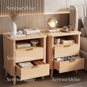 May include: Two light-toned wooden nightstands with vertical fluted side panels and four tapered legs. Each unit features two drawers with gold-toned rectangular handles. The tops hold books, a white vase with dried branches, and a spherical lamp.