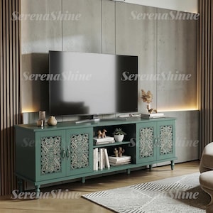 May include: A long, dark teal media console featuring four cabinet doors with intricate, carved floral patterns and silver-toned hardware. The unit includes an open central shelving section containing books and a small potted plant beneath a flat-screen TV.