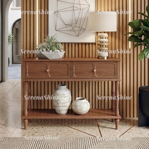 May include: A wooden console table with two drawers and a lower shelf, featuring a light-colored table lamp with a stacked stone base, a potted succulent in a geometric planter, and two textured ceramic vases on the bottom shelf.
