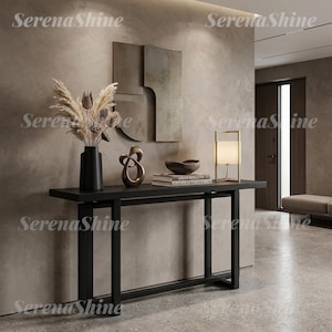 May include: A black console table with a rectangular top and four legs. On the table sits a black vase with dried pampas grass, a gold abstract sculpture, a dark bowl on two stacked books, and a gold-framed lamp with a white cylindrical shade.