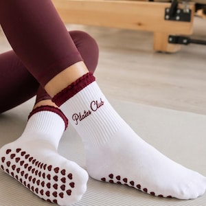 Pode incluir: Meias brancas com textura canelada e punho franzido em vermelho escuro. O texto "Pilates Club" está bordado em letra cursiva vermelha escura na lateral. As solas apresentam um padrão de pequenos pontos de aderência em silicone em forma de coração.