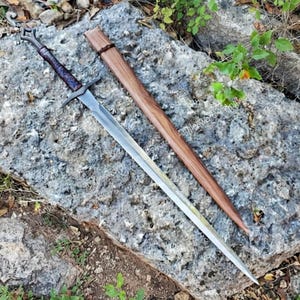 Può includere: Una spada in acciaio con impugnatura scura testurizzata e guardia, posizionata accanto a un fodero in legno abbinato. La lama presenta un solco centrale e una punta affilata, mentre il fodero mostra una venatura del legno naturale con una fascia scura.