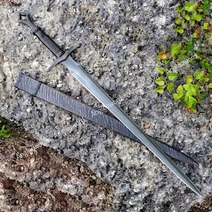 Op de afbeelding: Een donkergrijs metalen zwaard met een omwikkeld handvat en een ruitvormige pommel, vergezeld van een bijpassende gestructureerde schede. Het lemmet heeft een centrale richel en een punt, met de tekst Anglesword zichtbaar op lemmet en schede.