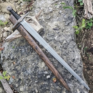 May include: A double-edged steel sword with a central fuller, featuring a black cord-wrapped hilt and a rounded pommel. The sword is partially inserted into a wooden scabbard with a distinct dark brown and black wood grain pattern.