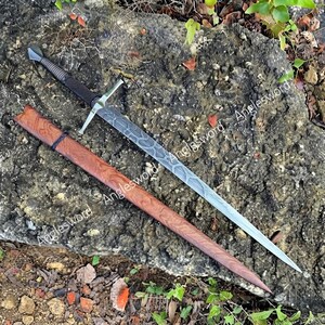 May include: A longsword with a double-edged steel blade featuring a complex etched pattern, a silver-toned crossguard, and a black cord-wrapped handle. The sword is accompanied by a wooden scabbard with a distinct reddish-brown wood grain finish.