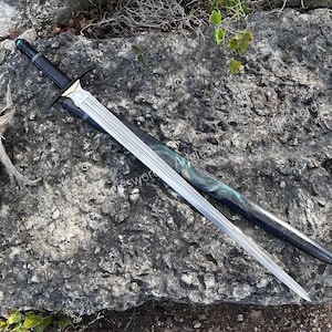 Può includere: Una spada lunga con lama in argento metallizzato dotata di scanalatura centrale, impugnatura nera con pietra blu sul pomolo e guardia color oro. La spada è accompagnata da un fodero nero con un motivo vorticoso blu e verde.