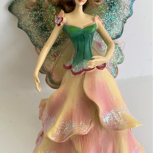 May include: A fairy figurine with a green bodice and a petal-like skirt in shades of yellow and pink. The fairy has glittery green wings and long, light brown hair. The figurine is a decorative item.